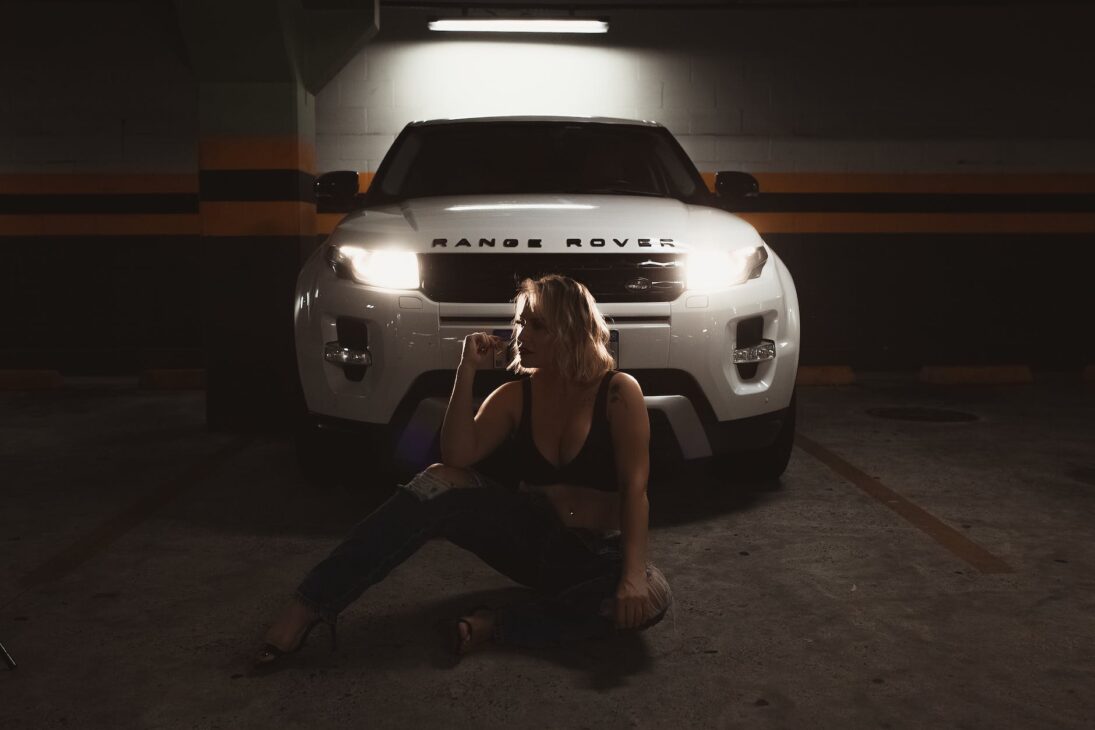 woman wearing a black bra sitting on a garage floor and posing with a white vehicle